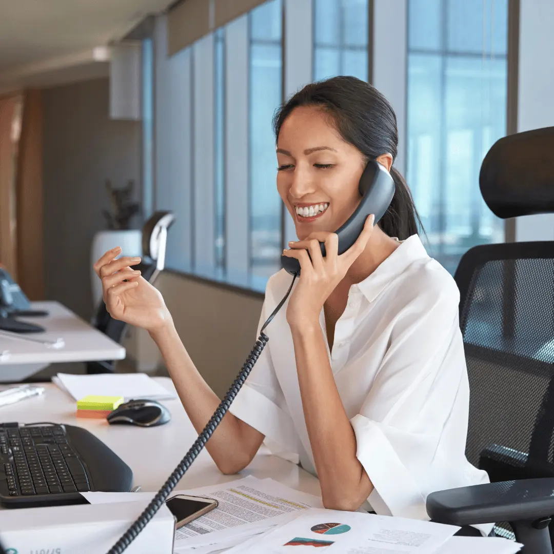 Woman in a white blouse sits at a desk, smiling while talking on a corded office phone in a bright workspace.
