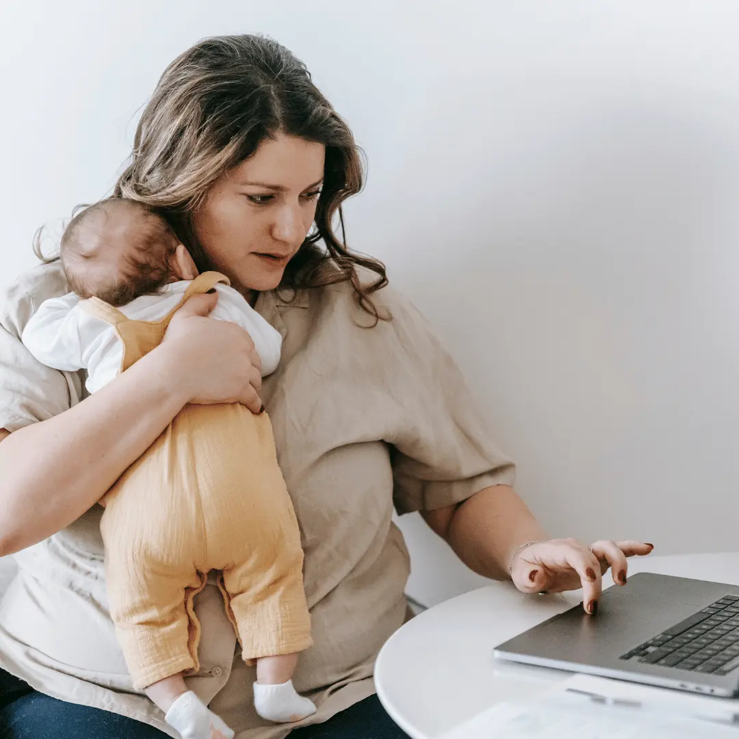 Mother sits at a round white table, holding a baby in a mustard carrier while using a laptop nearby.